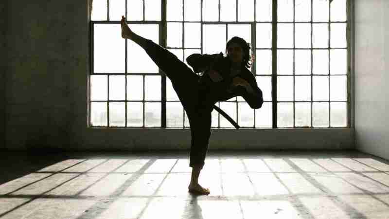 Martial artist performing a high kick in a sunlit room creating dramatic shadows., tags: und - pexel